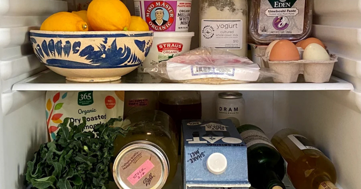 Close-up of a stocked refrigerator shelf with a bowl of lemons and fresh cilantro in view, alongside eggs, milk, and jarred goods