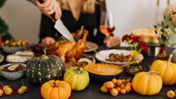 Hands carving a roasted turkey at a festive fall dinner table decorated with pumpkins, seasonal fruits, flowers, and autumn-themed dishes.