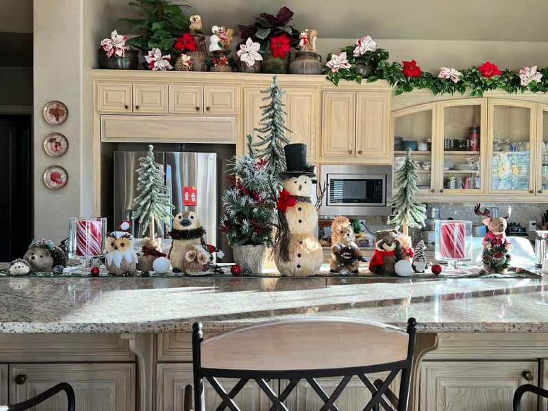 A kitchen counter is adorned with an array of festive decorations including whimsical snowmen, forest animals, and a garland, with cabinet tops featuring poinsettias and white flowers, radiating holiday cheer.