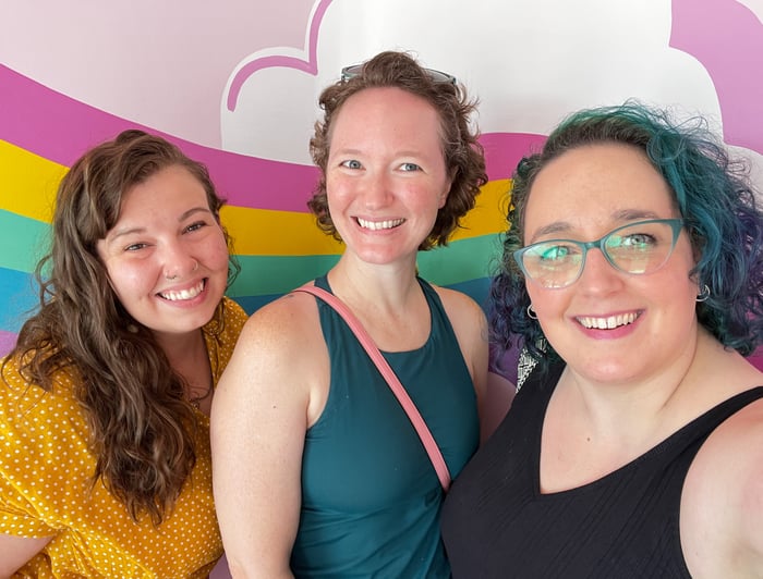 A bright and colorful photo of three women smiling in front of a pastel rainbow. Megan is on the left, Kate in the middle and Heather on the right.