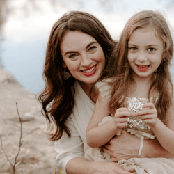 A smiling photo of a woman with long, wavy brown hair with her smiling young daughter. They're sitting on a rock in front of a stream.