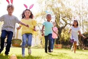 Children wearing bunny ears running across a grassy yard with Easter baskets during an egg hunt