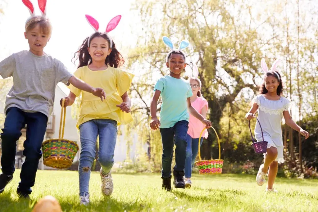 Children wearing bunny ears running across a grassy yard with Easter baskets during an egg hunt